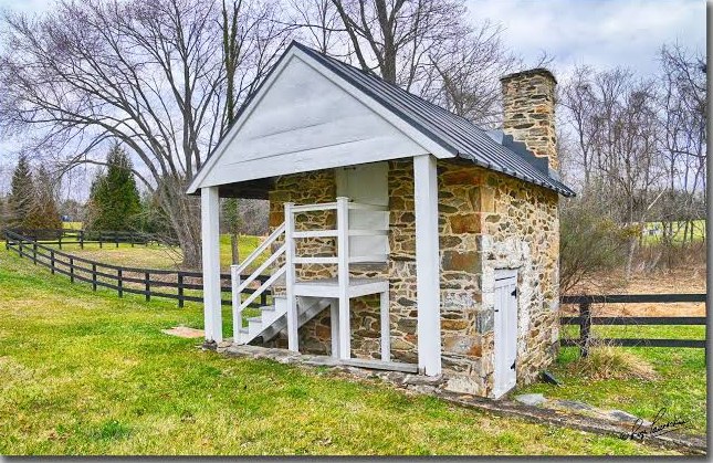 A fieldstone springhouse with a white porch and stairs, beside a split rail fence